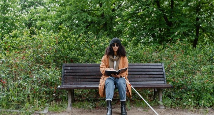 a tan skinned Black woman sitting on a park bench and reading she has a white cane used by visuallu imparied people.jpg.optimal