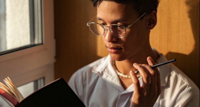 an Asian man with lightly tanned skin holding a pencil and reading a book.jpg.optimal