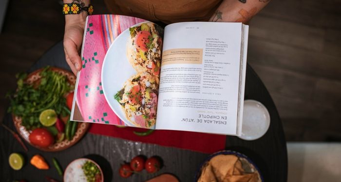 a medium light skinned person with tattoes holding open a cookbook in a kitchen.jpg.optimal