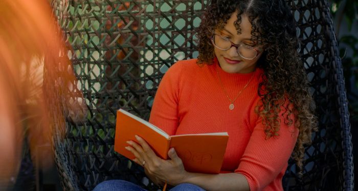 a tanned skin woman of color reading a book in a woven chair.jpg.optimal