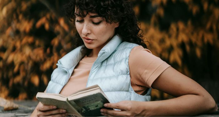 a lightly tanned skin woman reading outside with fall foliage in the background.jpg.optimal