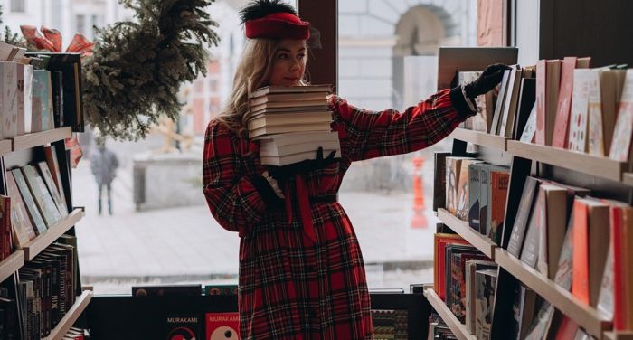 a white woman in red plaid with a stack of books reaching for another book on the shelf in a bookstore.jpg.optimal