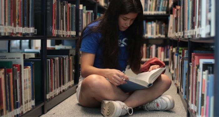 a medium light skinned young woman sits cross legged on library floor while reading a book.jpg.optimal