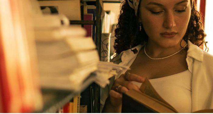 fair skinned woman with curly hair and a scarf reading a book against a bookshelf.jpg.optimal